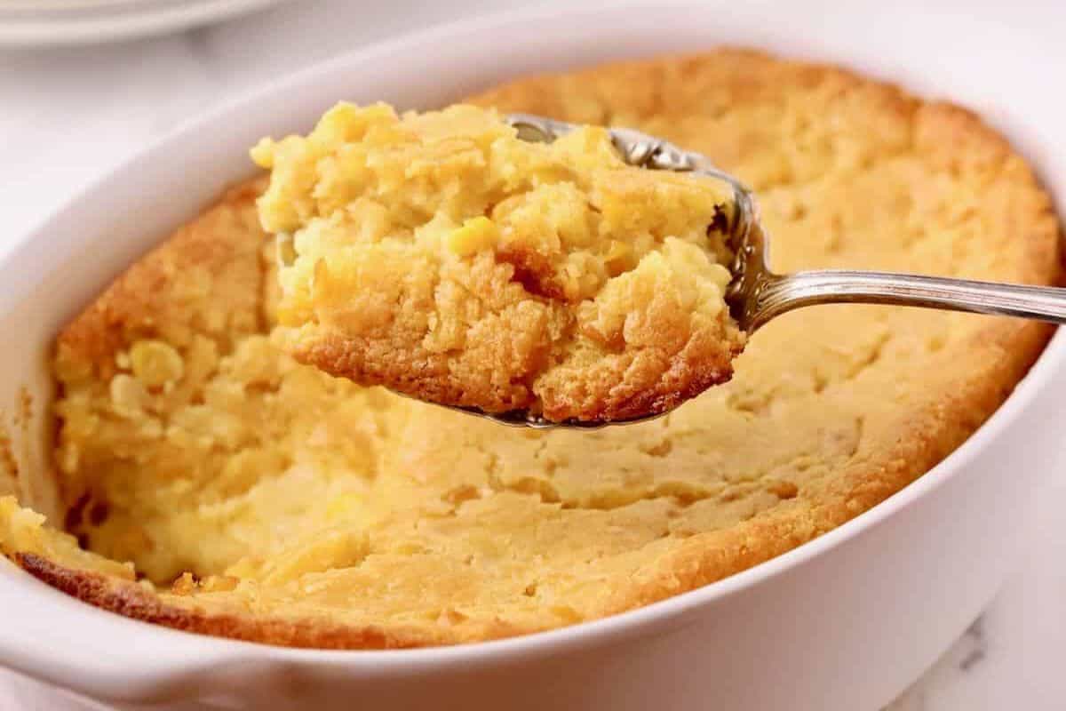 A spoon scoops a portion of baked corn casserole from a white baking dish, making it an ideal addition to your Easter side dishes.