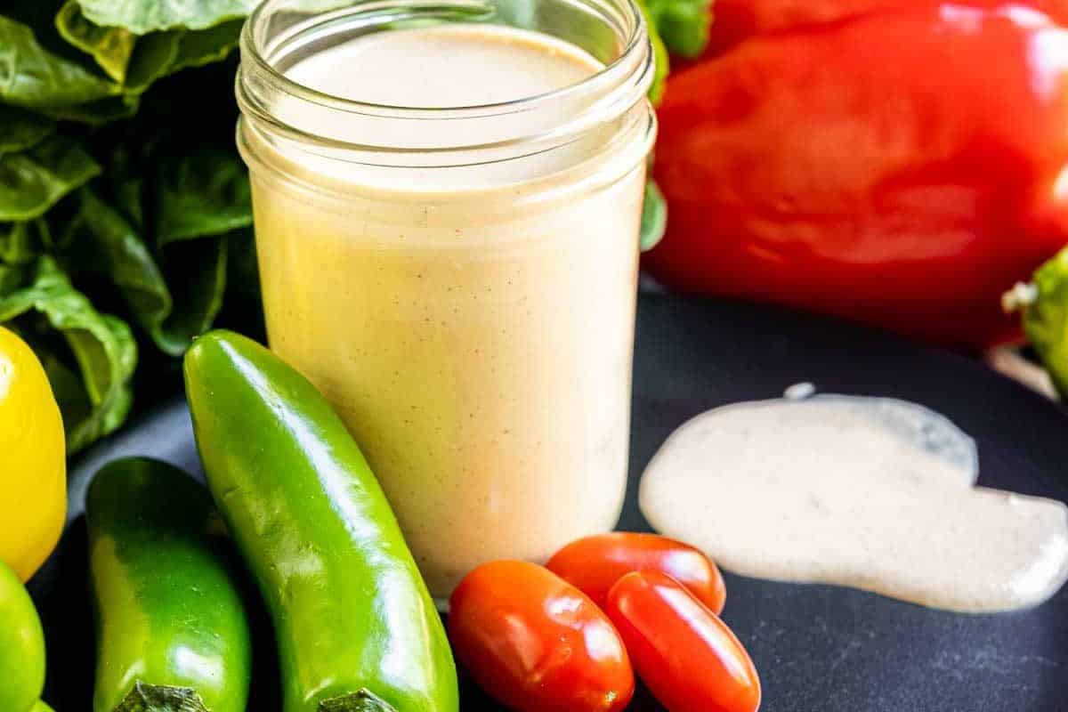 Southwest salad dressing in a glass jar, surrounded by jalapeños, cherry tomatoes, and a red bell pepper on a black plate.