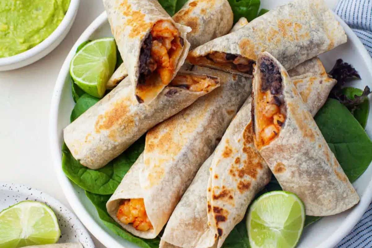 A plate of homemade burritos brimming with beans and a rich red-orange filling, served on spinach leaves with lime wedges. In the background, a bowl of guacamole adds a festive touch, perfect for celebrating Cinco de Mayo.
