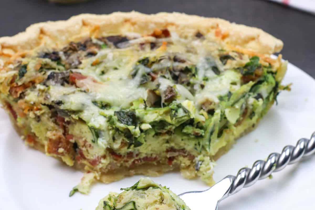 Slice of spinch and bacon quiche on a white plate with a fork, showcasing a delightful mix of greens, cheese, and a flaky crust—perfect for your spring brunch recipes.
