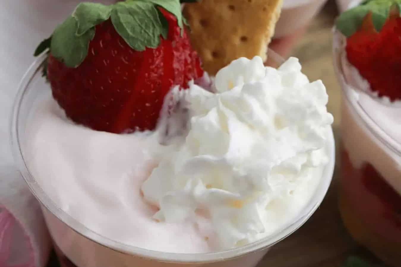 A beautiful dessert cup with whipped cream, a whole strawberry adorned with green leaves, and a piece of graham cracker on top showcases the essence of spring desserts.