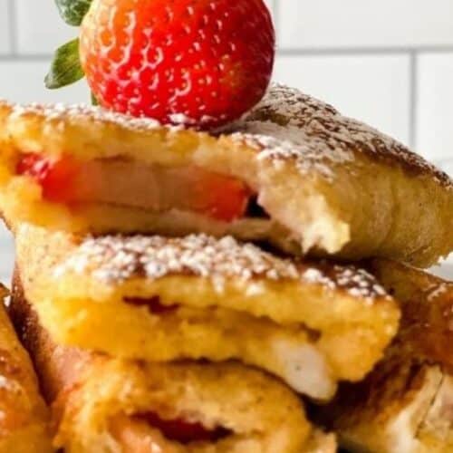 Close-up of three stacked French toast rolls with strawberry filling, dusted with powdered sugar, and topped with a whole strawberry against a tiled background. Perfect to impress at spring brunch gatherings.