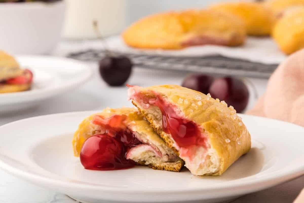 A cherry-filled pastry with a golden crust is sliced open, revealing its luscious filling. Fresh cherries dot the background on a cooling rack and plate, making this dessert perfect for any festive occasion like Cinco de Mayo.