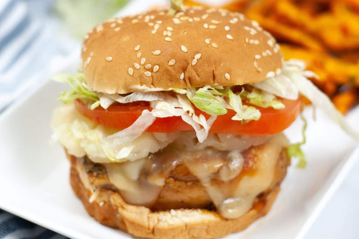 Teriyaki chicken burger with melted cheese, tomato, and lettuce on a sesame bun, served on a white plate with sweet potato fries in the background.