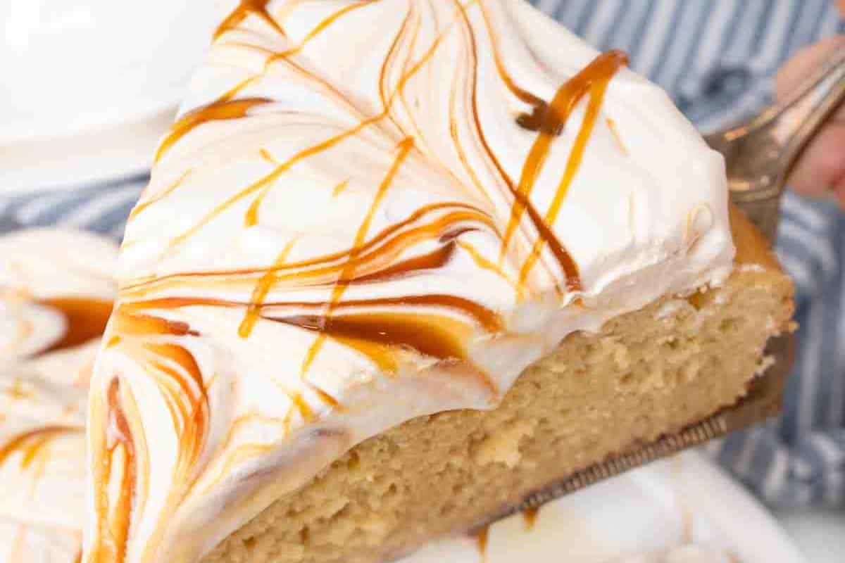 A slice of tres leches cake, perfect for a Cinco de Mayo fiesta, is topped with white frosting and caramel drizzle, being lifted gracefully on a spatula.