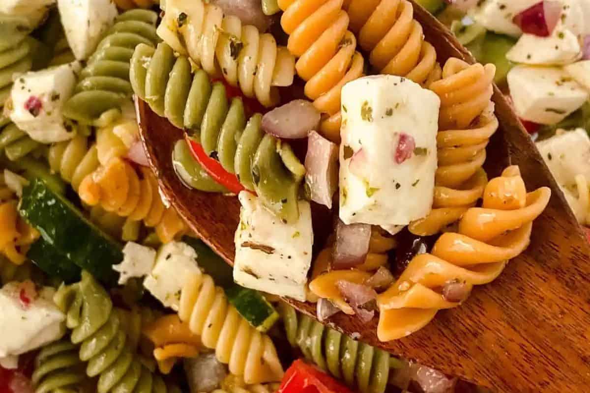 A wooden spoon scoops up a vibrant rotini pasta salad featuring tricolor pasta, feta cheese, diced cucumbers, tomatoes, and onions—an inviting dish perfect for those seeking the best spring salads.
