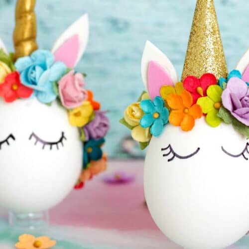 Two eggs decorated as unicorns with golden horns, flower crowns, and painted closed eyes are showcased as a delightful Easter egg decorating idea, set against a soft colorful background.