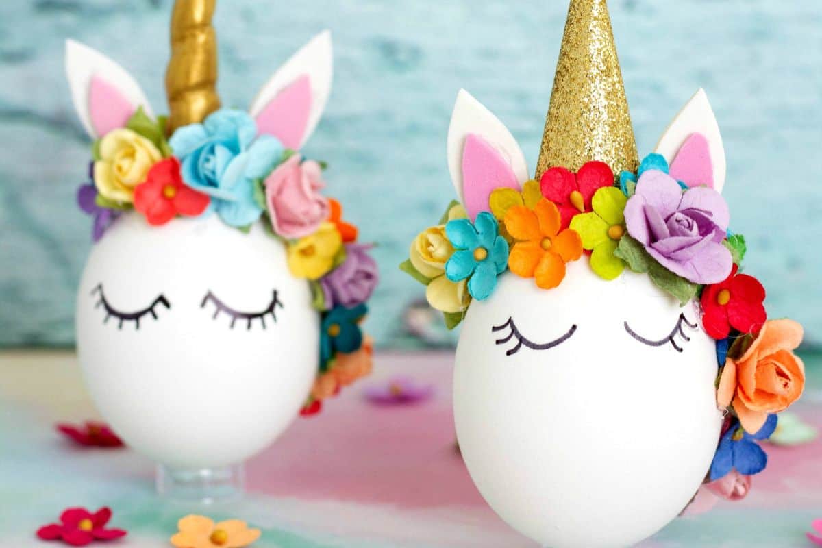 Two eggs decorated as unicorns with golden horns, flower crowns, and painted closed eyes are showcased as a delightful Easter egg decorating idea, set against a soft colorful background.