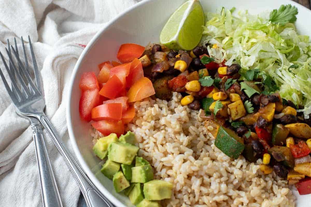 A vibrant bowl featuring brown rice, diced tomatoes, avocado, lettuce, black beans, corn, zucchini, and bell peppers. Perfect for a Cinco de Mayo celebration or as a refreshing twist on homemade burritos. Garnished with lime wedges and two inviting forks nearby.
