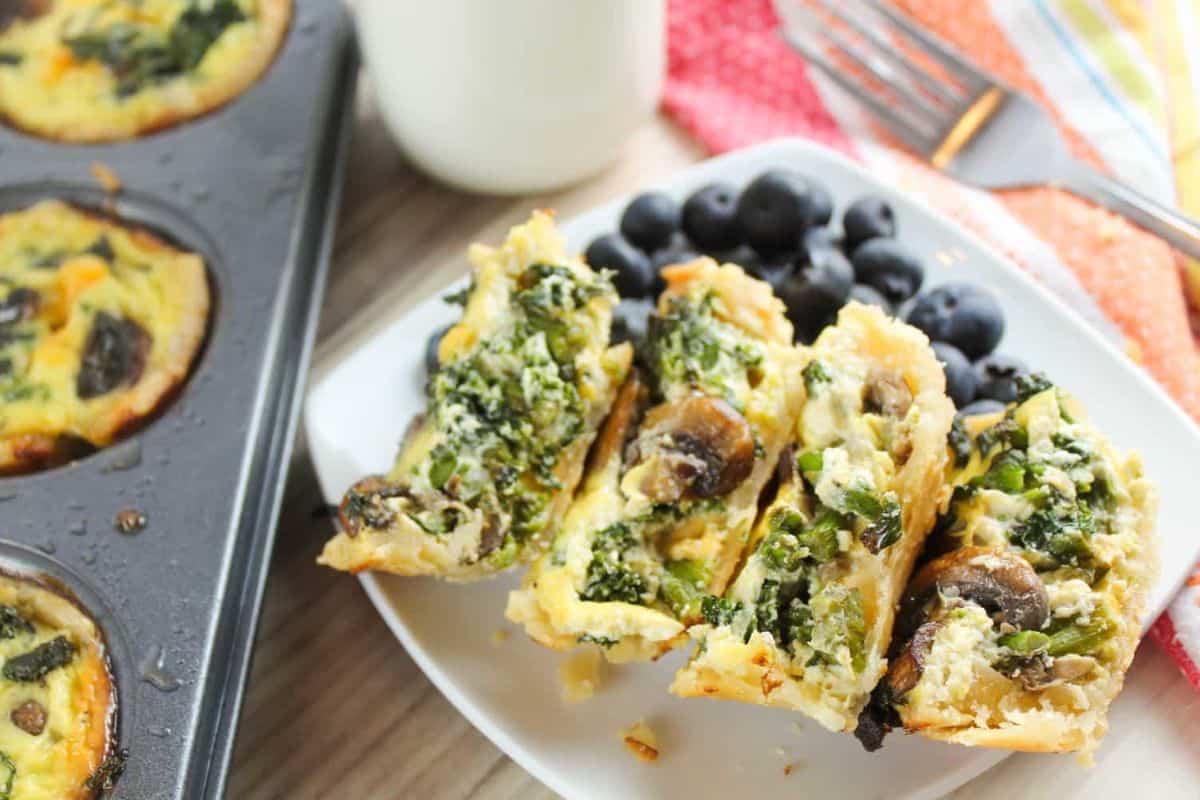 Mini quiches with spinach and mushrooms grace a plate beside fresh blueberries, embodying delightful spring brunch recipes. In the background, a muffin tray sits alongside a glass of milk and a striped cloth, setting the scene for a perfect morning gathering.