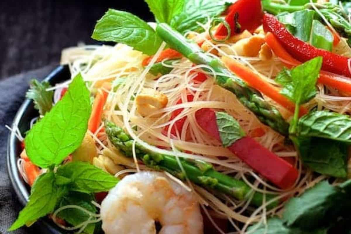 A vibrant dish features vermicelli noodles tossed with shrimp, asparagus, red bell peppers, and carrots, all garnished with fresh mint leaves—truly one of the best spring salad recipes.