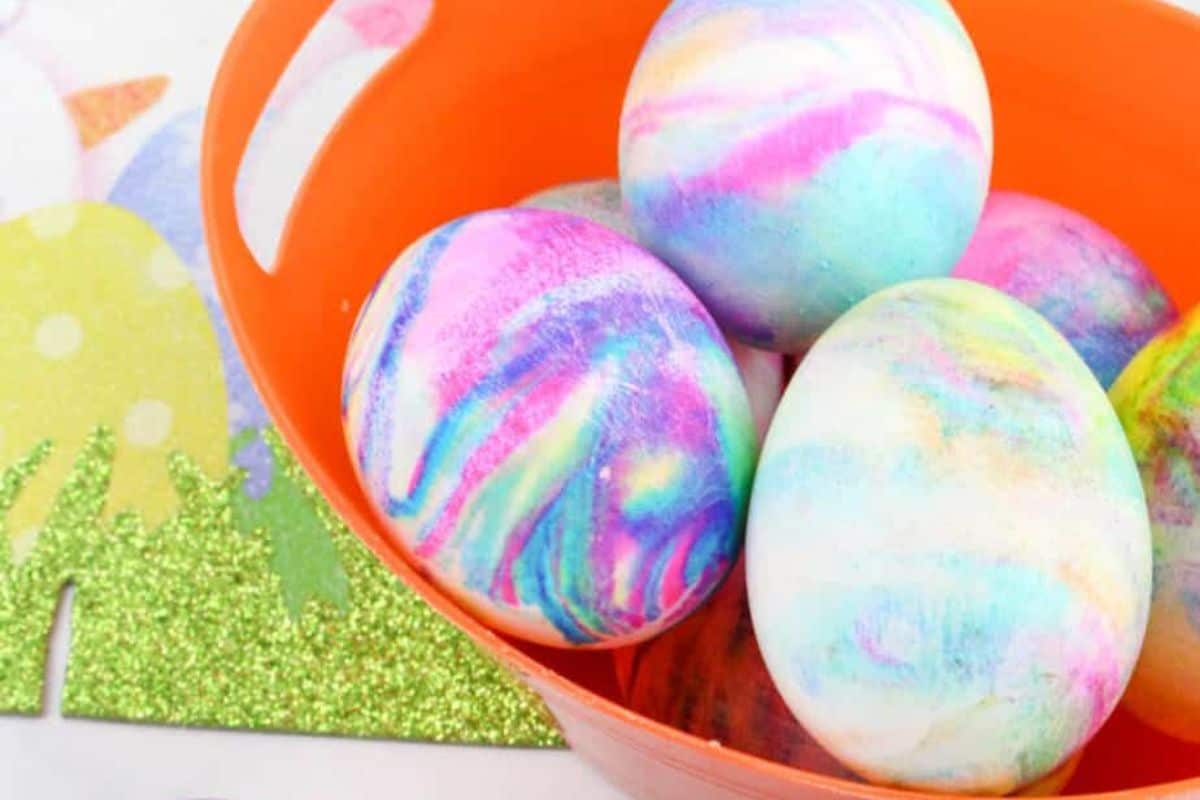 Colorfully dyed Easter eggs in a vibrant orange bowl showcase creative ideas for Easter egg decorating, placed on a decorative surface with grass and yellow shapes.