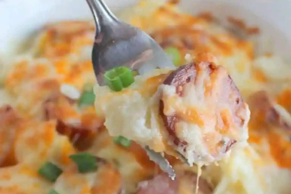 A fork holds a piece of sausage with melted cheese and green onions, hovering above a dish of creamy baked casserole, evoking cravings for irresistible dishes.