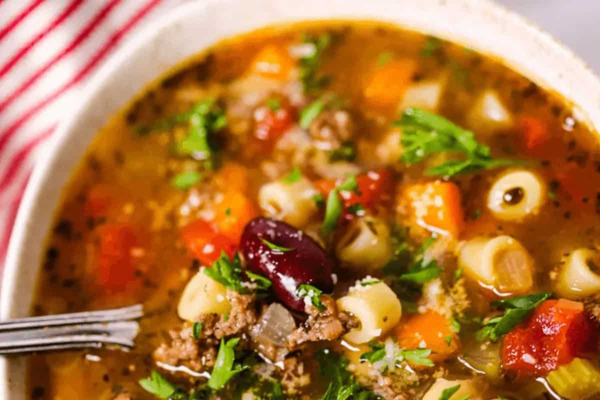 A flavorful bowl of vegetable and pasta soup features tube-shaped pasta, kidney beans, diced tomatoes, carrots, and ground meat. Garnished with fresh parsley for a delightful touch, this dish is perfect for those seeking lent-friendly pasta recipes. A spoon rests inside the bowl.