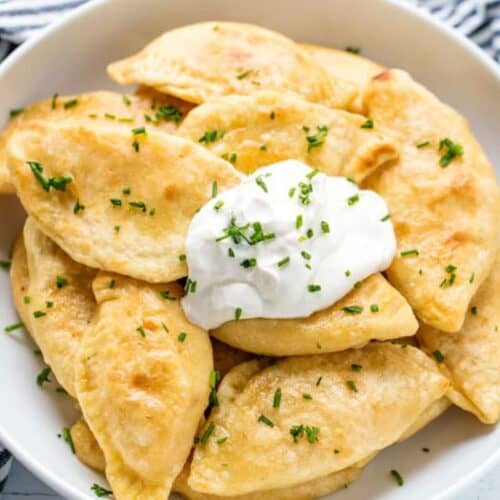 A plate of golden-brown pierogi, an irresistible delight topped with a dollop of sour cream and sprinkled with chopped herbs, tempts your cravings. A striped cloth is seen partially in the background.