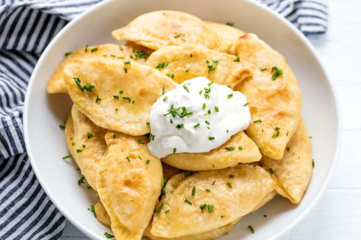 A plate of golden-brown pierogi, an irresistible delight topped with a dollop of sour cream and sprinkled with chopped herbs, tempts your cravings. A striped cloth is seen partially in the background.
