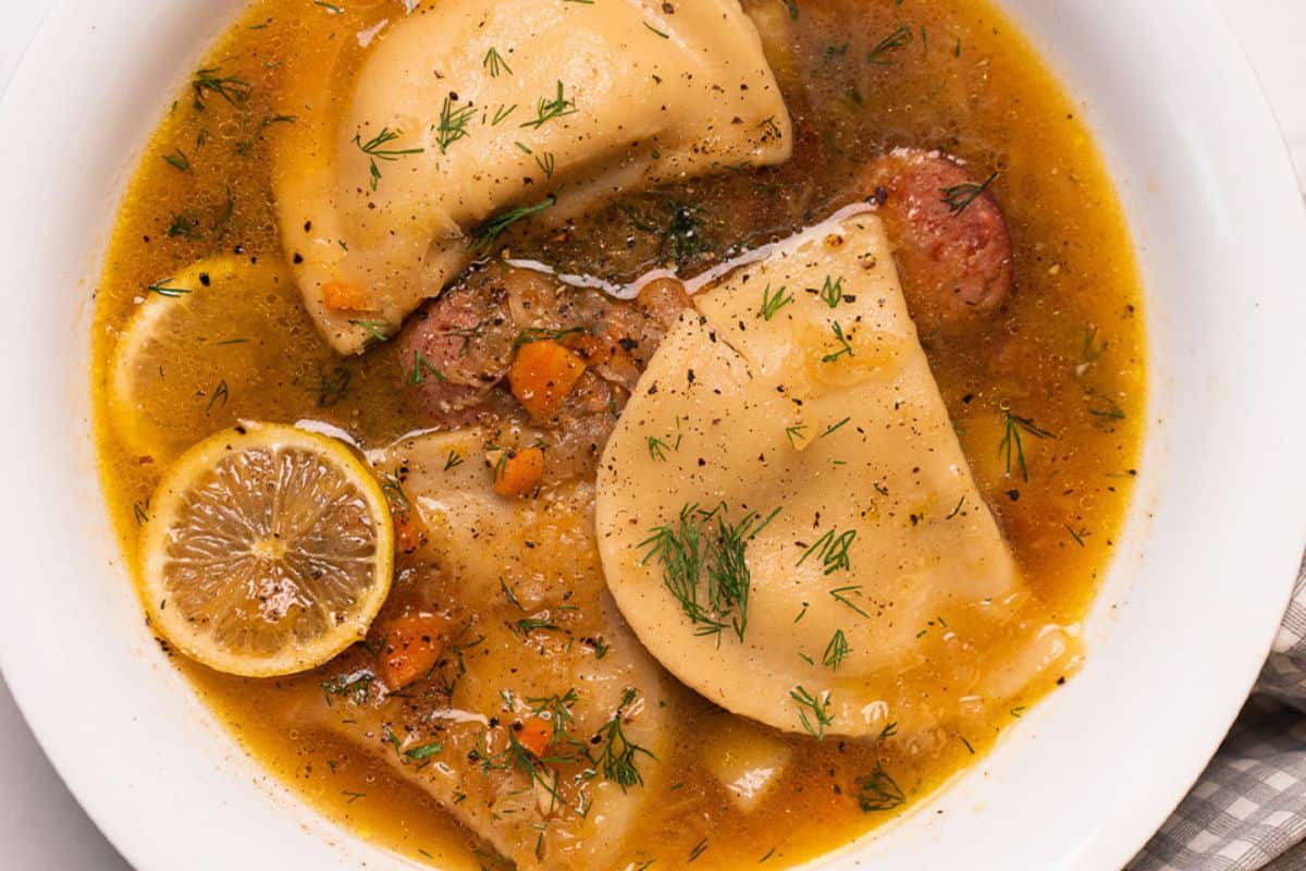 Satisfy your cravings with a comforting soup featuring hearty dumplings, tangy sliced lemon, fresh carrots, herbs, and savory sausage all cozily nestled in a white bowl.
