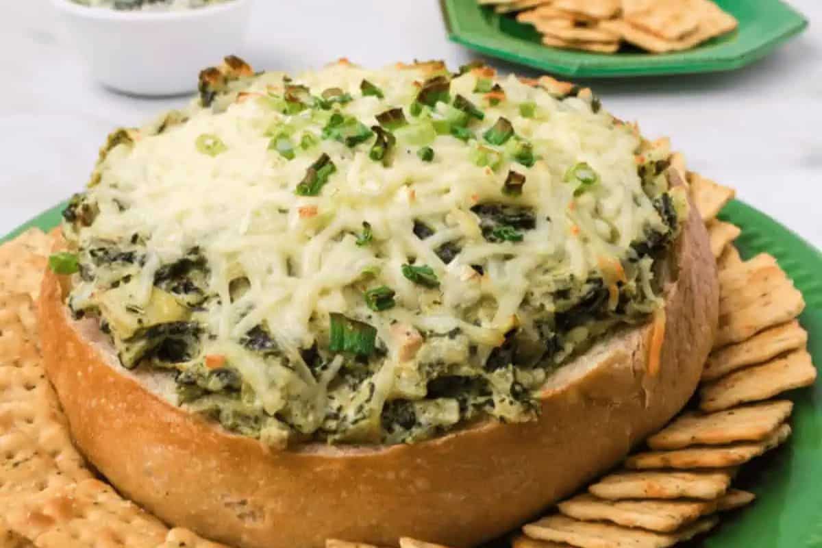 A round bread bowl filled with cheesy spinach artichoke dip, topped with chopped green onions, surrounded by crackers on a green plate—perfect for Lent and delicious meatless recipes.