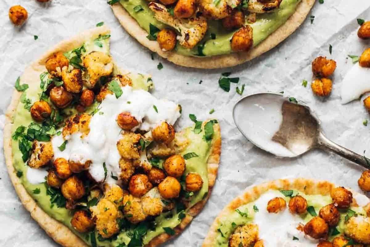Pita bread topped with mashed avocado, roasted chickpeas, cauliflower, chopped herbs, and a drizzle of white sauce—a perfect addition to your collection of delicious Lent recipes, served with a spoon on parchment paper.