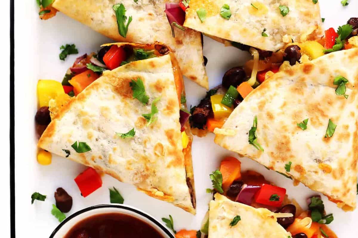 Sliced quesadilla with melted cheese, black beans, and colorful diced vegetables, garnished with chopped cilantro, served next to a small bowl of dipping sauce—perfect for Lent or anyone seeking delicious meatless recipes.
