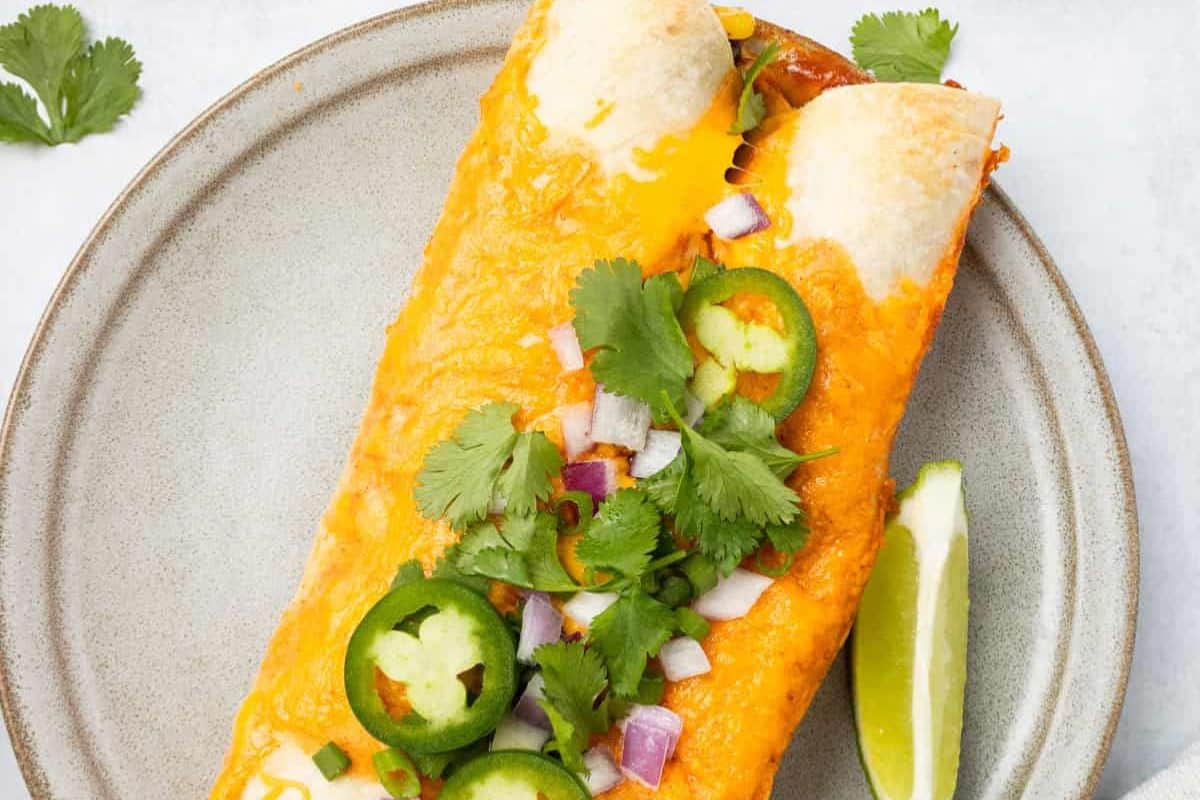 A cheese-covered enchilada topped with sliced jalapeños, chopped red onion, and cilantro, served with a lime wedge on a beige plate—perfect for Lent or anyone seeking delicious meatless recipes.