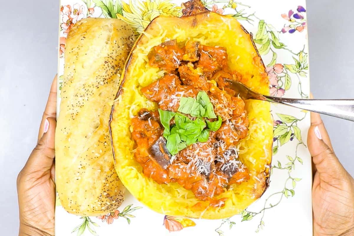 A person holds a floral plate with spaghetti squash filled with tomato sauce and herbs, topped with grated cheese, next to a seeded bread roll—perfect for Lent and those seeking meatless meals.