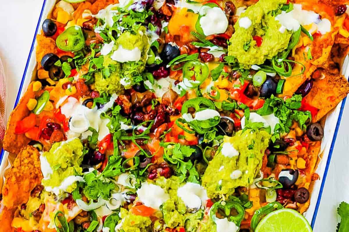 A tray of loaded nachos topped with guacamole, sour cream, chopped vegetables, sliced olives, scallions, cilantro, and lime wedges—perfect for meatless recipes or as a delicious vegetarian Lent meal.