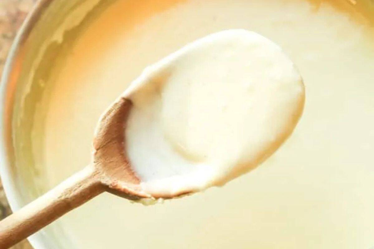 A close-up of a wooden spoon holding a generous amount of creamy, pale white Alfredo Sauce over a bowl filled with more of the sauce.
