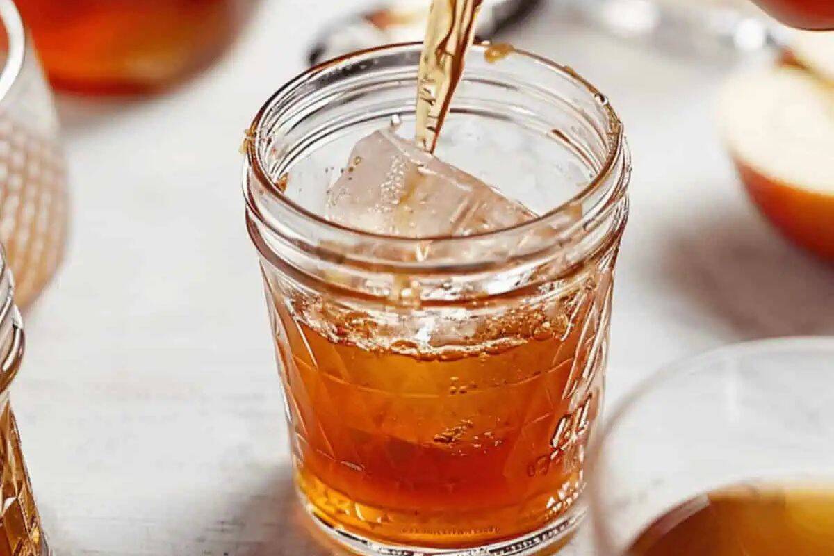 A close-up shot of a clear glass mason jar being filled with amber-colored Apple Pie Moonshine. An ice cube is visible in the jar.