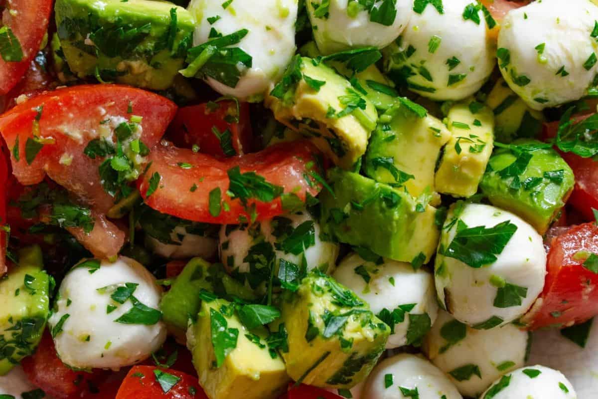 A close-up overhead shot of Avocado Mozzarella Tomato Salad. Chunks of creamy green avocado are mixed with small white mozzarella balls and pieces of ripe red tomatoes, all tossed with fresh green herbs.