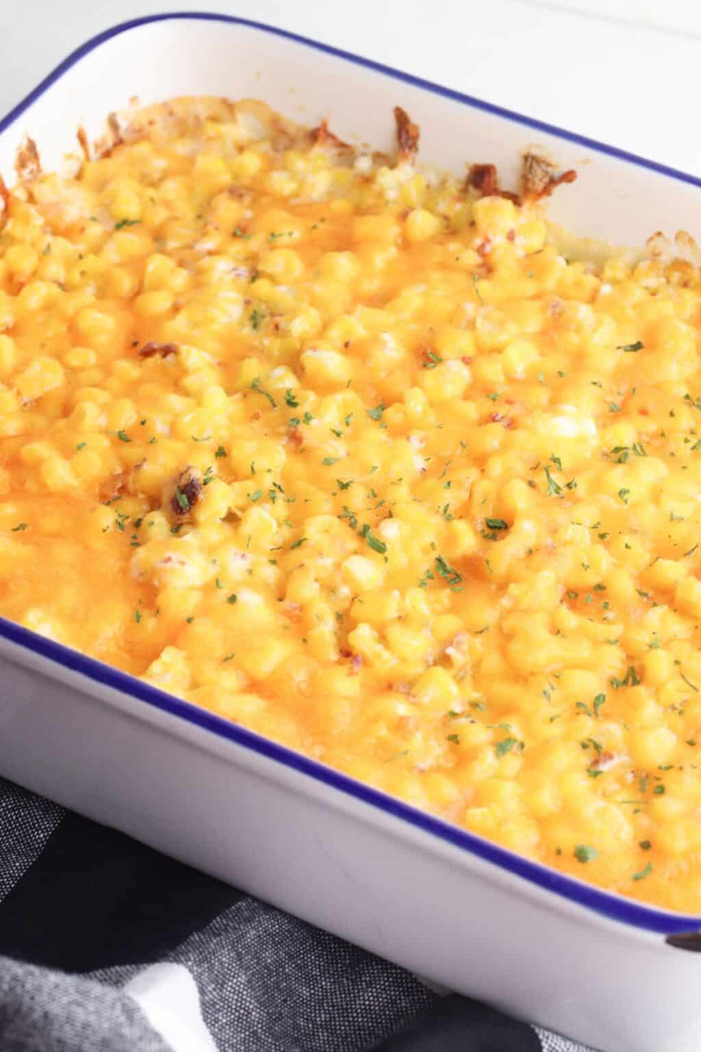 Corn Casserole with Cream Cheese: Baked casserole dish filled with creamy corn and rich cream cheese, topped with melted cheese and herbs, resting on a black and white cloth.