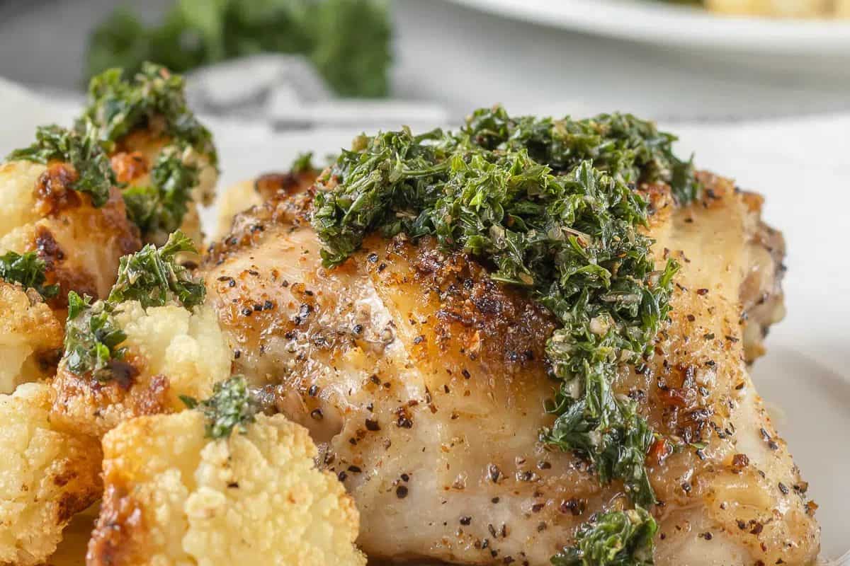 Roasted chicken thigh topped with chopped herbs, served with pieces of roasted cauliflower on a white plate—a perfect addition to your collection of juicy chicken thigh recipes.