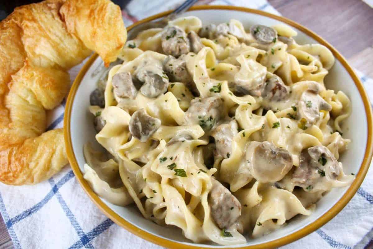 Wide egg noodles are coated in a creamy, light brown sauce with tender strips of beef and sliced mushrooms, garnished with chopped fresh parsley. A croissant is visible on the side.