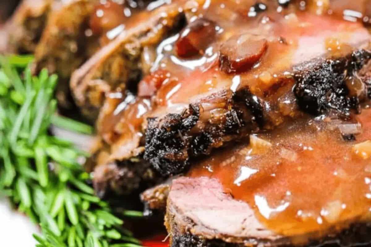  A close-up shot of sliced beef tenderloin, showcasing its pink center, generously coated with a glossy, dark brown port wine sauce. Fresh rosemary sprigs are visible on the side.