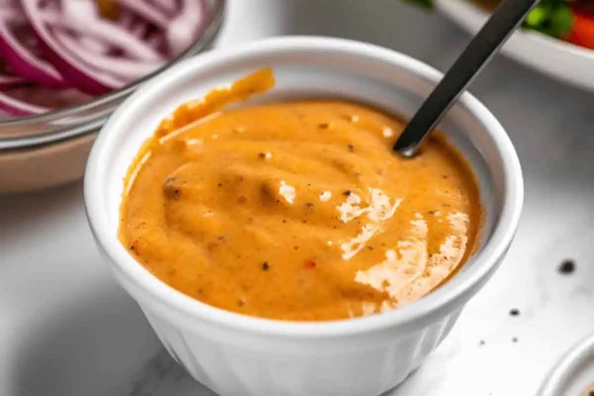 A close-up overhead shot of a small white ramekin filled with creamy, pale orange-pink Best Burger Sauce. A black spoon rests inside the sauce, and small flecks of spices or relish are visible.