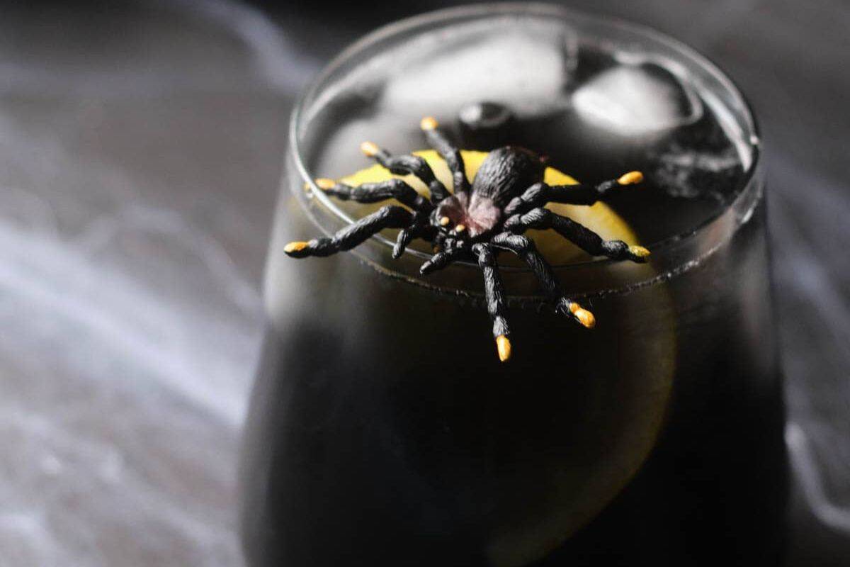 A black drink with ice and a lemon slice in a glass, topped with a realistic plastic spider decoration—this spooky twist puts a fun spin on timeless cocktails everyone should know.