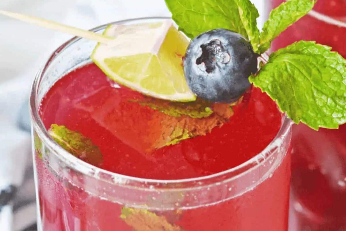 A close-up shot of a vibrant red Blueberry Mojito in a clear glass. The drink is garnished with fresh mint sprigs, a juicy blueberry, and a lime wedge on a skewer.