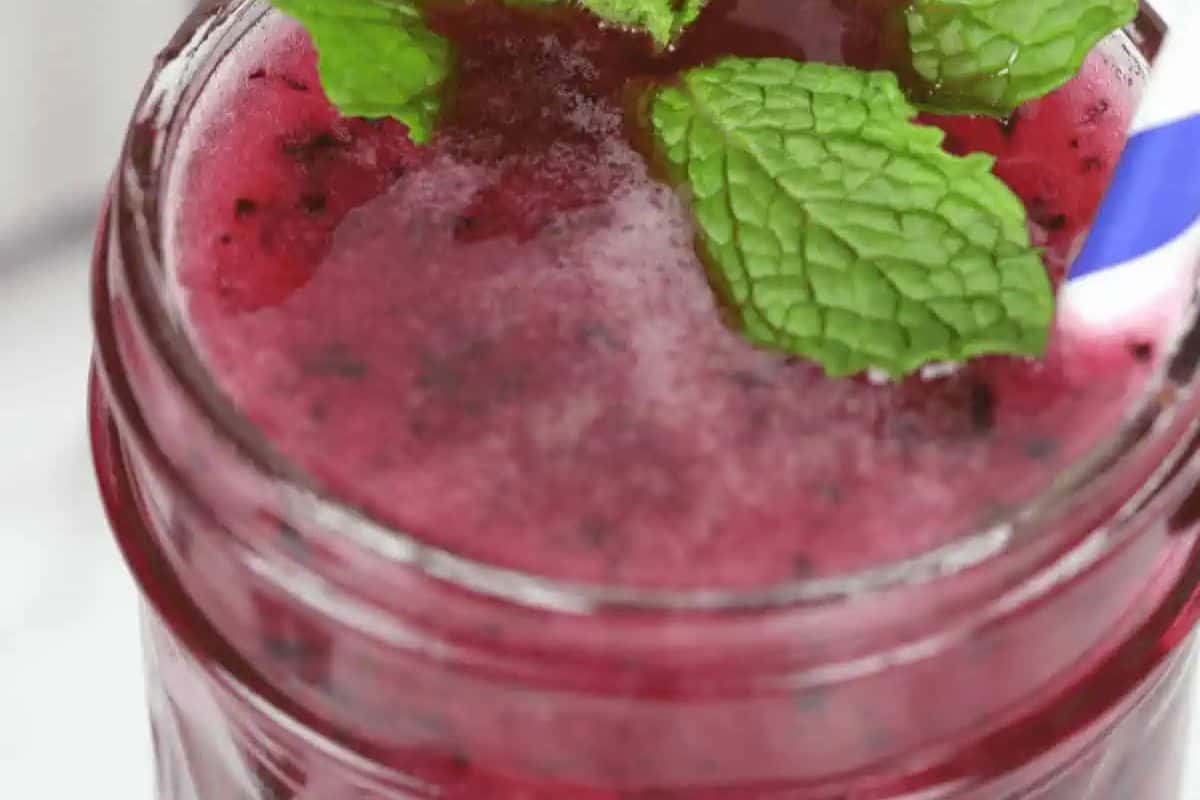 A close-up of a glass jar filled with a vibrant red Blueberry Moonshine. It is garnished with fresh mint sprigs and a blue and white striped straw.
