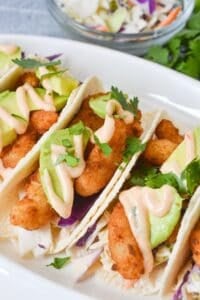 Three fish tacos with avocado slices, shredded cabbage, cilantro, and a drizzle of creamy sauce are arranged on a white plate. Reminiscent of popular shrimp tacos like Boom Boom Shrimp Tacos, these flavorful bites are perfect for any taco lover. A bowl of slaw is visible in the background.