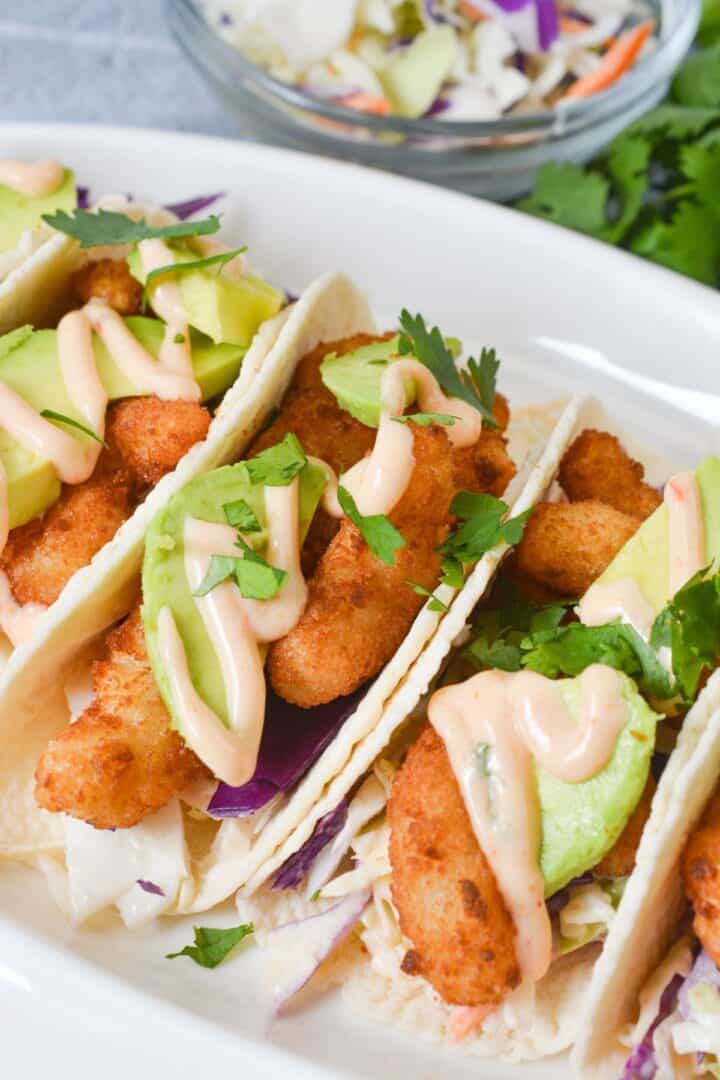 Three fish tacos with avocado slices, shredded cabbage, cilantro, and a drizzle of creamy sauce are arranged on a white plate. Reminiscent of popular shrimp tacos like Boom Boom Shrimp Tacos, these flavorful bites are perfect for any taco lover. A bowl of slaw is visible in the background.