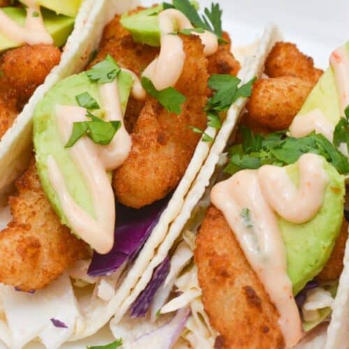 Three fish tacos with battered fish, avocado slices, shredded cabbage, creamy sauce, and chopped cilantro on soft tortillas, arranged on a white plate—perfect for pairing with our signature Boom Boom Shrimp Tacos.