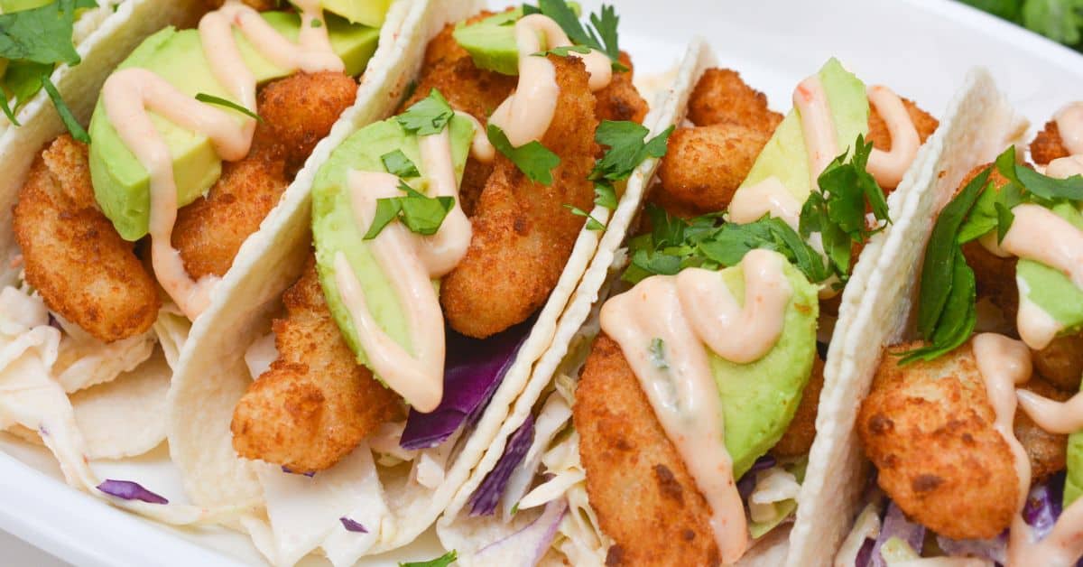 Three fish tacos with battered fish, avocado slices, shredded cabbage, creamy sauce, and chopped cilantro on soft tortillas, arranged on a white plate—perfect for pairing with our signature Boom Boom Shrimp Tacos.