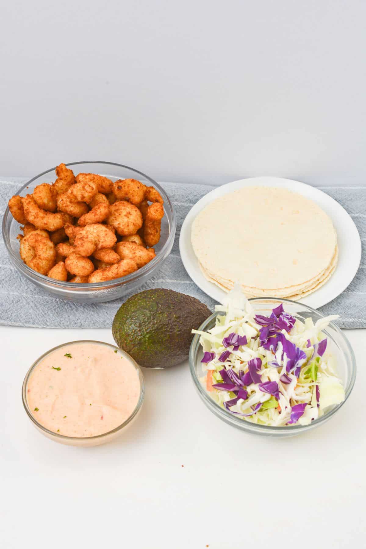 Four bowls contain battered shrimp, tortillas, shredded cabbage, and a creamy sauce; an avocado sits between them on a white surface, ready to be transformed into delicious Boom Boom Shrimp Tacos.