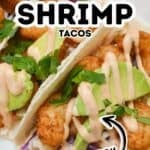 Boom Boom Shrimp Tacos feature crispy shrimp tacos topped with avocado slices, chopped cilantro, shredded cabbage, and a drizzle of creamy sauce, all served on a white plate.