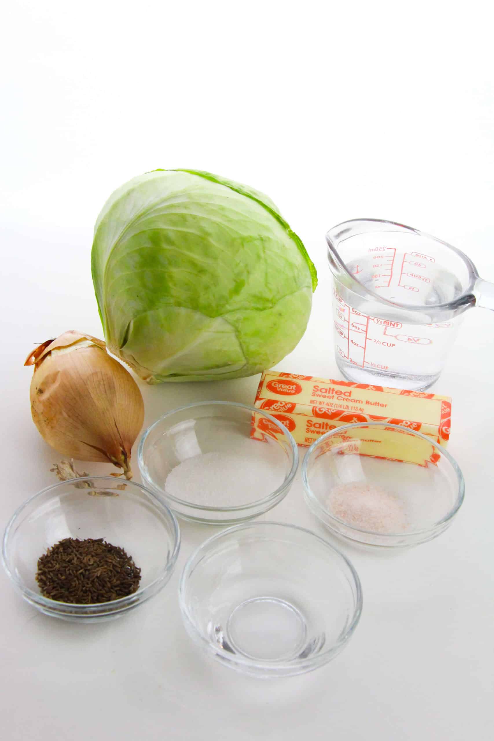 Ingredients for the dish on a white surface: a head of the vegetables, an onion, a stick of salted butter, a measuring cup of water, caraway seeds, sugar, salt in small bowls, and an empty bowl.