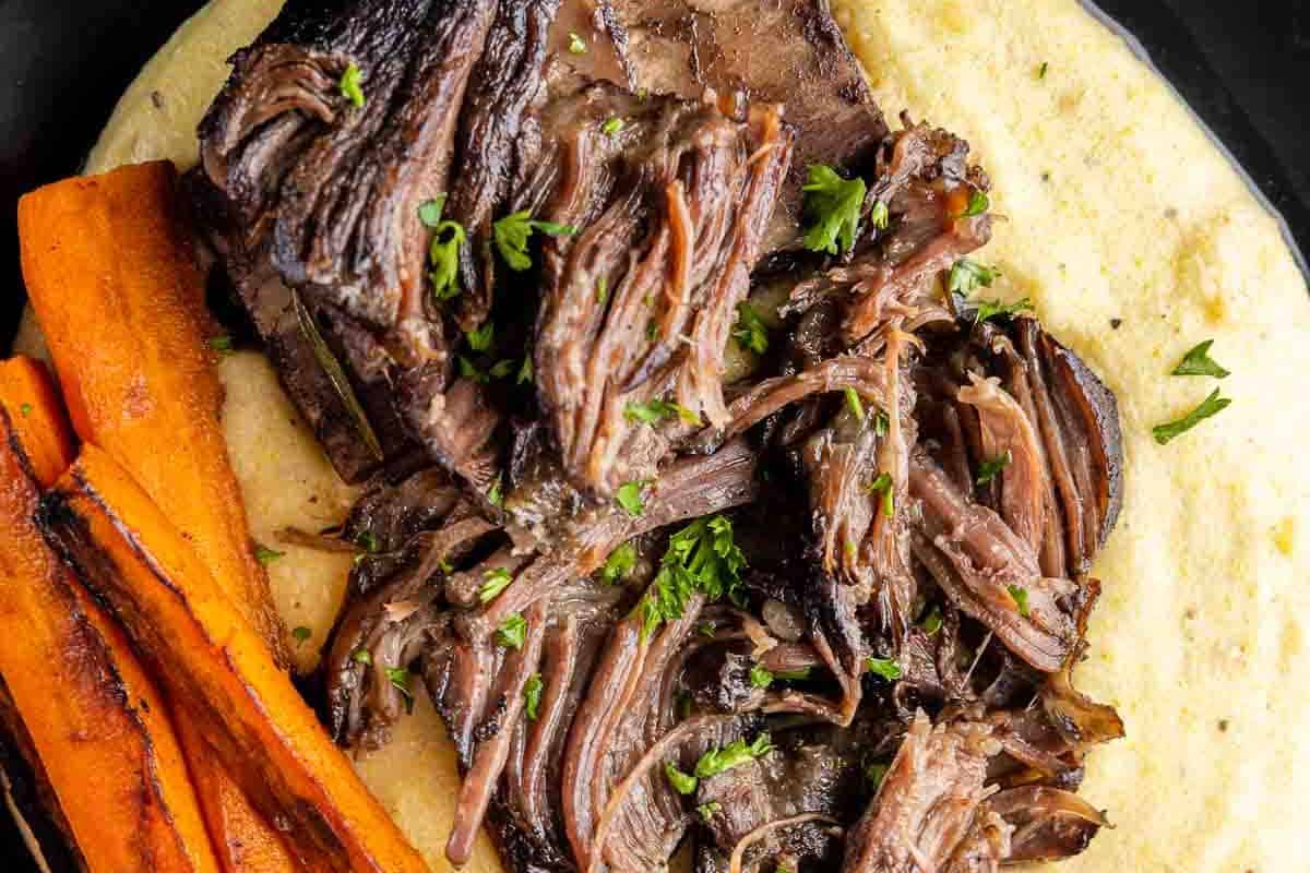 A close-up overhead shot of tender, shredded Braised Short Ribs served over a bed of creamy polenta or mashed potatoes, alongside glazed carrots.