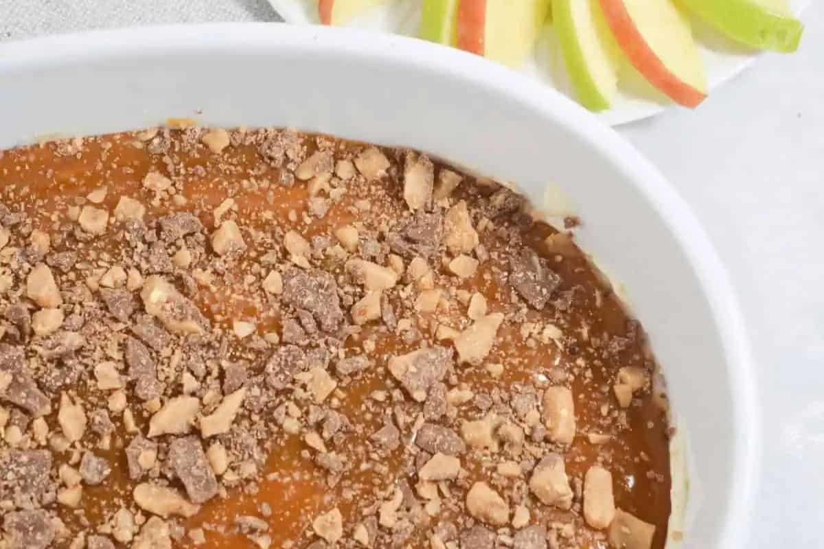 A white dish filled with easy caramel toffee-topped dip sits next to a plate with slices of green and red apples.