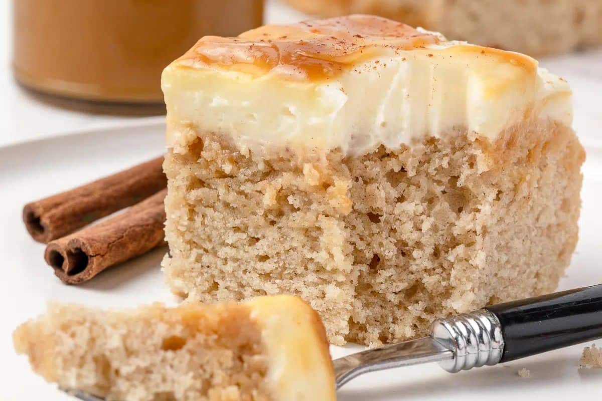 A square slice of cinnamon spice cake with creamy frosting on a white plate, next to two cinnamon sticks and a fork holding a bite of this indulgent dessert.