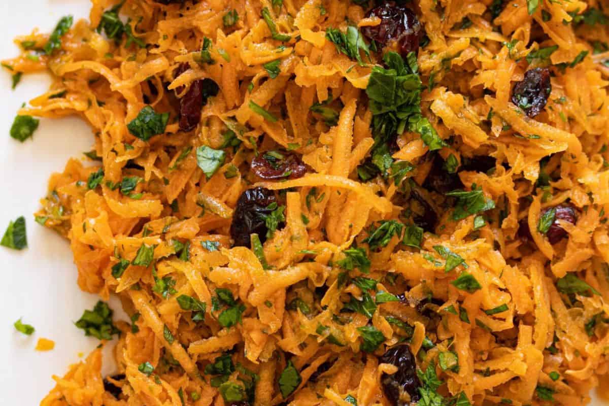 Close-up of a shredded carrot salad with chopped herbs and dark dried fruit pieces, possibly raisins or cranberries, mixed throughout—a nod to classic recipes that celebrate timeless cuisine.