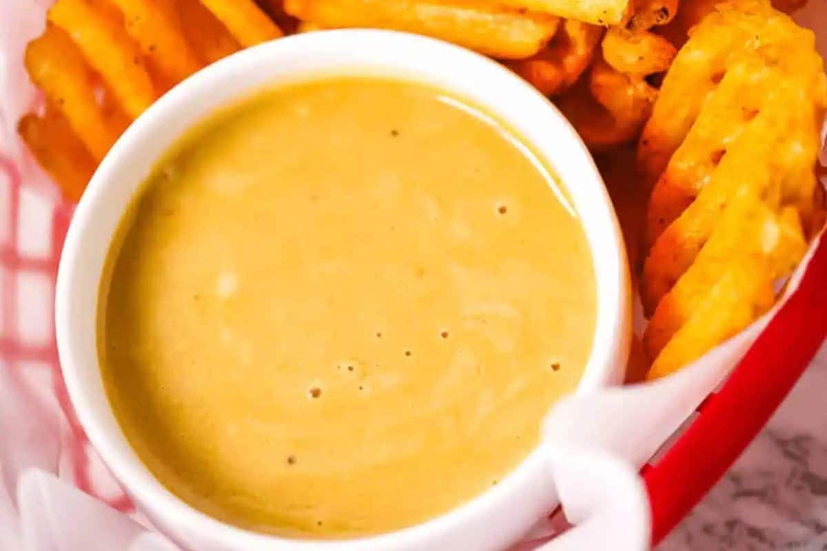 A close-up overhead shot of a small white bowl filled with creamy, light brown Chick-fil-A Sauce, nestled amongst golden-brown waffle fries in a red and white paper-lined basket.